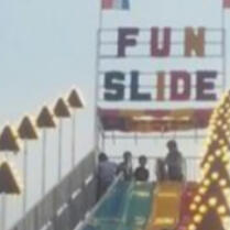 the top of a large slide, complete with a sign reading 'FUN SLIDE' in large, mutlicolor letters. Children are waiting at the top